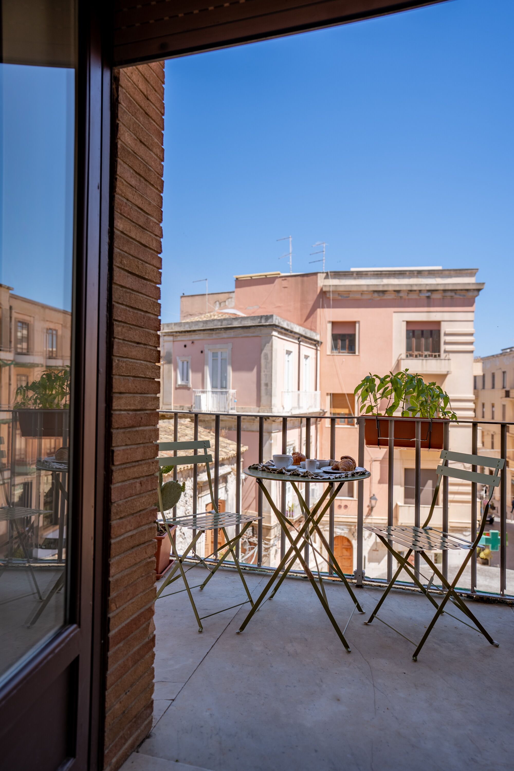 A balcony with a table and chairs, potted plants, and a view of neighboring buildings.