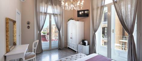 A hotel room with a bed, a desk, a chair, a wardrobe, a TV, and a balcony with outdoor seating.