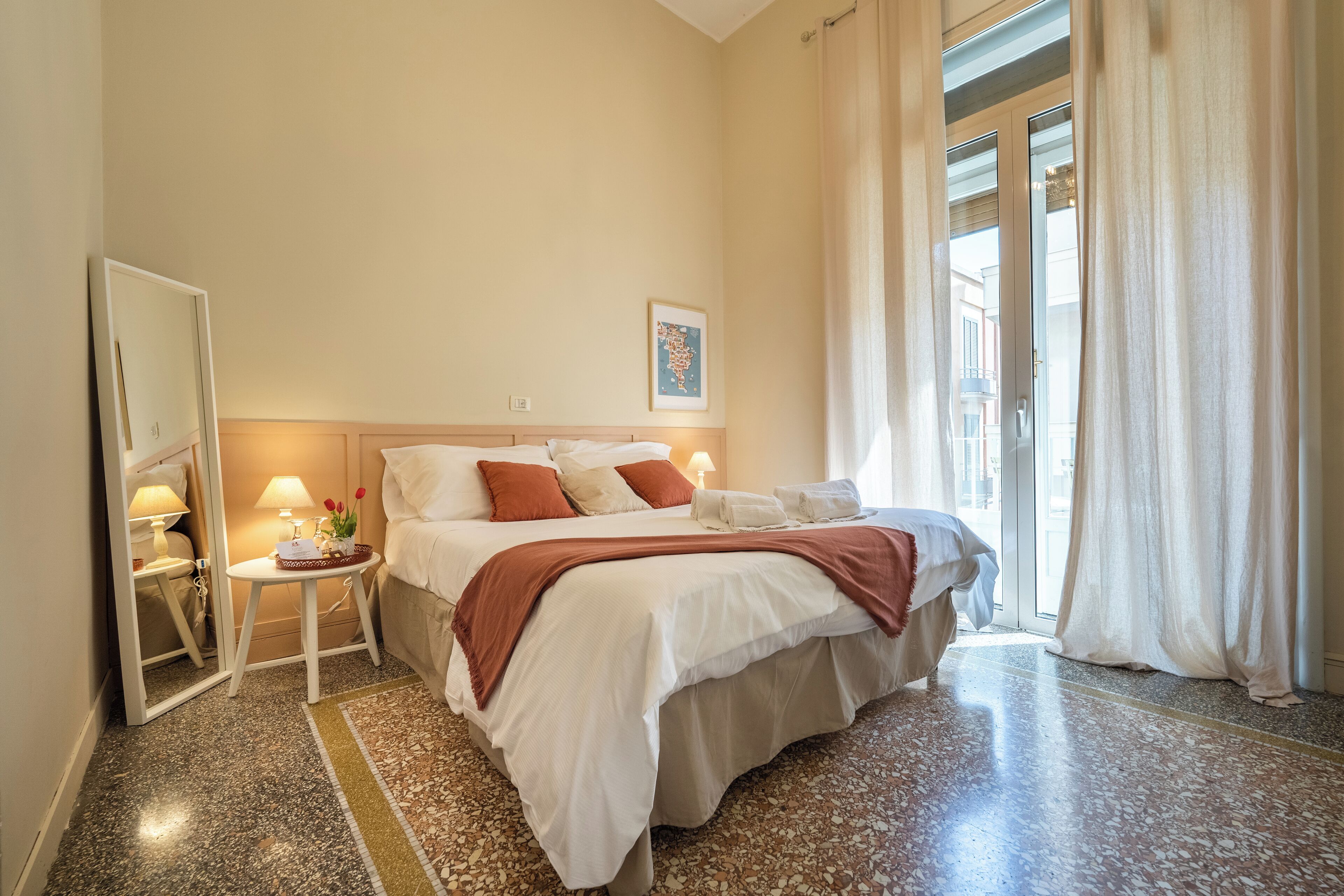 A hotel room with a large bed, a nightstand with a lamp, a mirror, and a window with curtains.