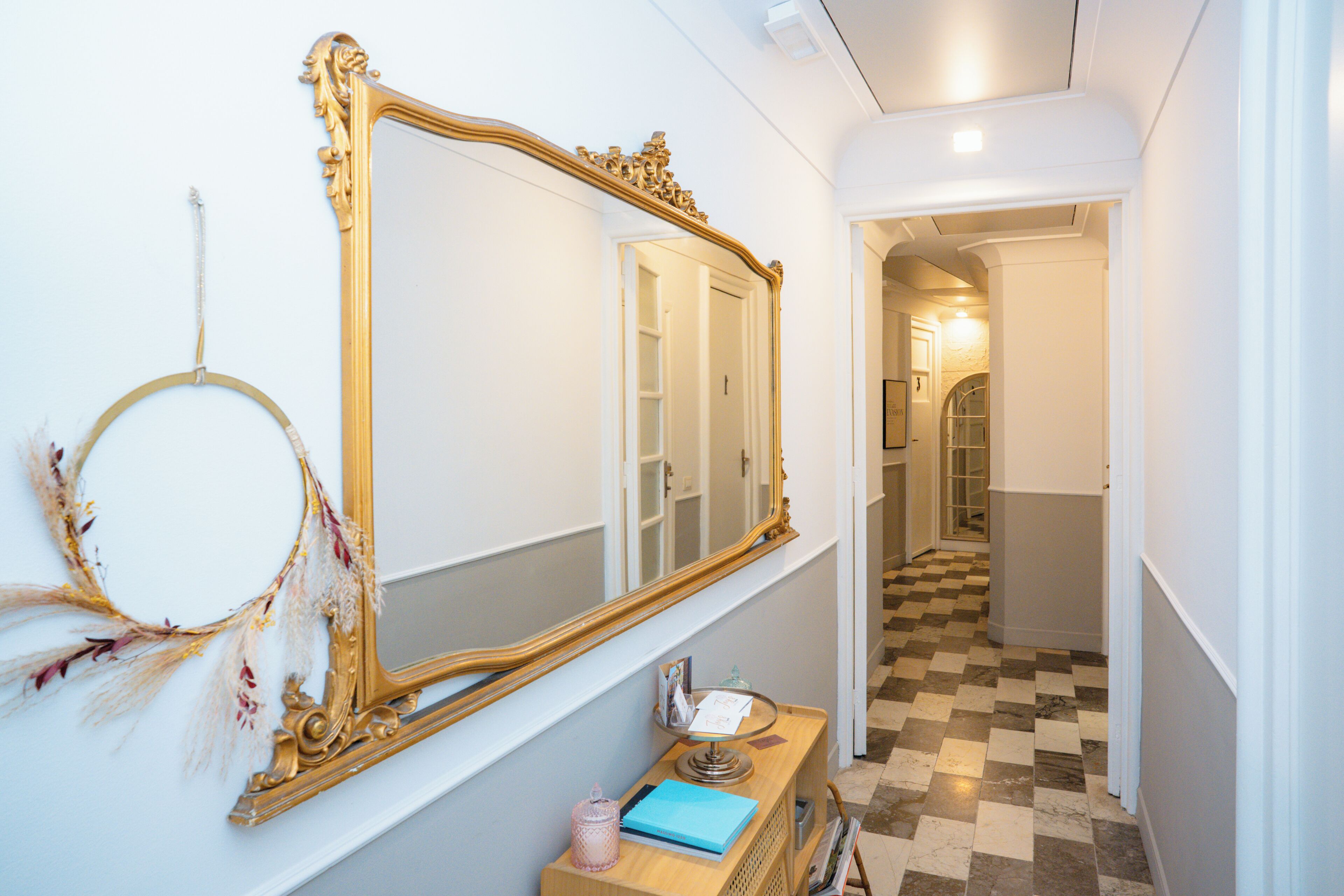 A hallway with a large ornate mirror, a small table with a book and a candle, and a checkered floor.
