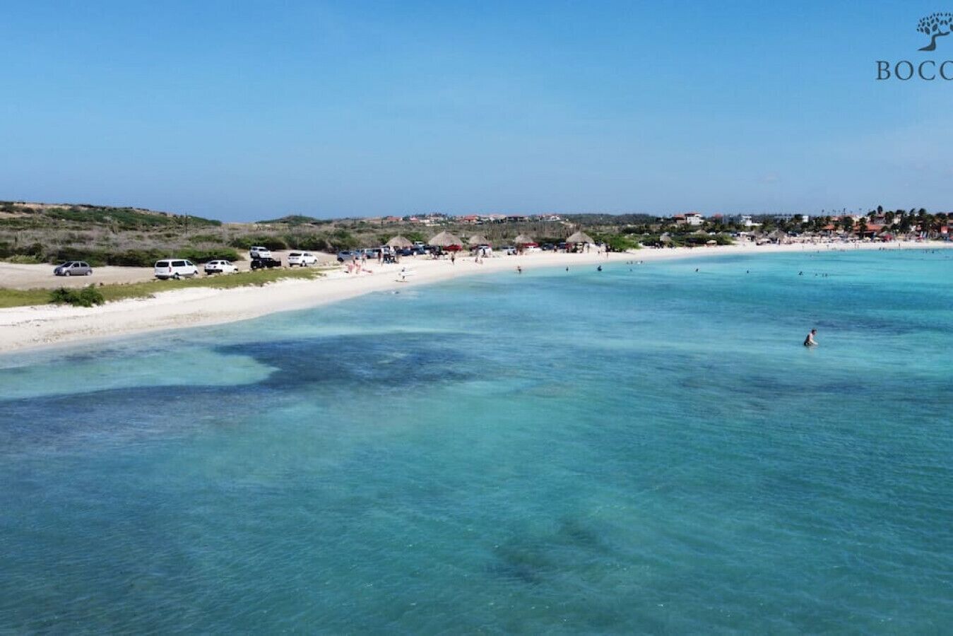 Une plage avec une eau turquoise et limpide, un rivage sablonneux et quelques bâtiments en arrière-plan.