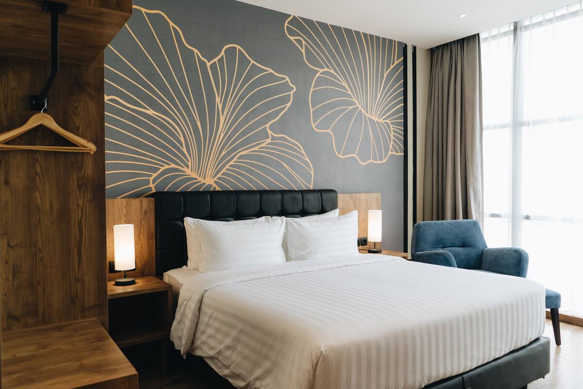 Une chambre d’hôtel moderne avec un grand lit, deux tables de chevet, un fauteuil bleu et un mur orné d’un motif floral.