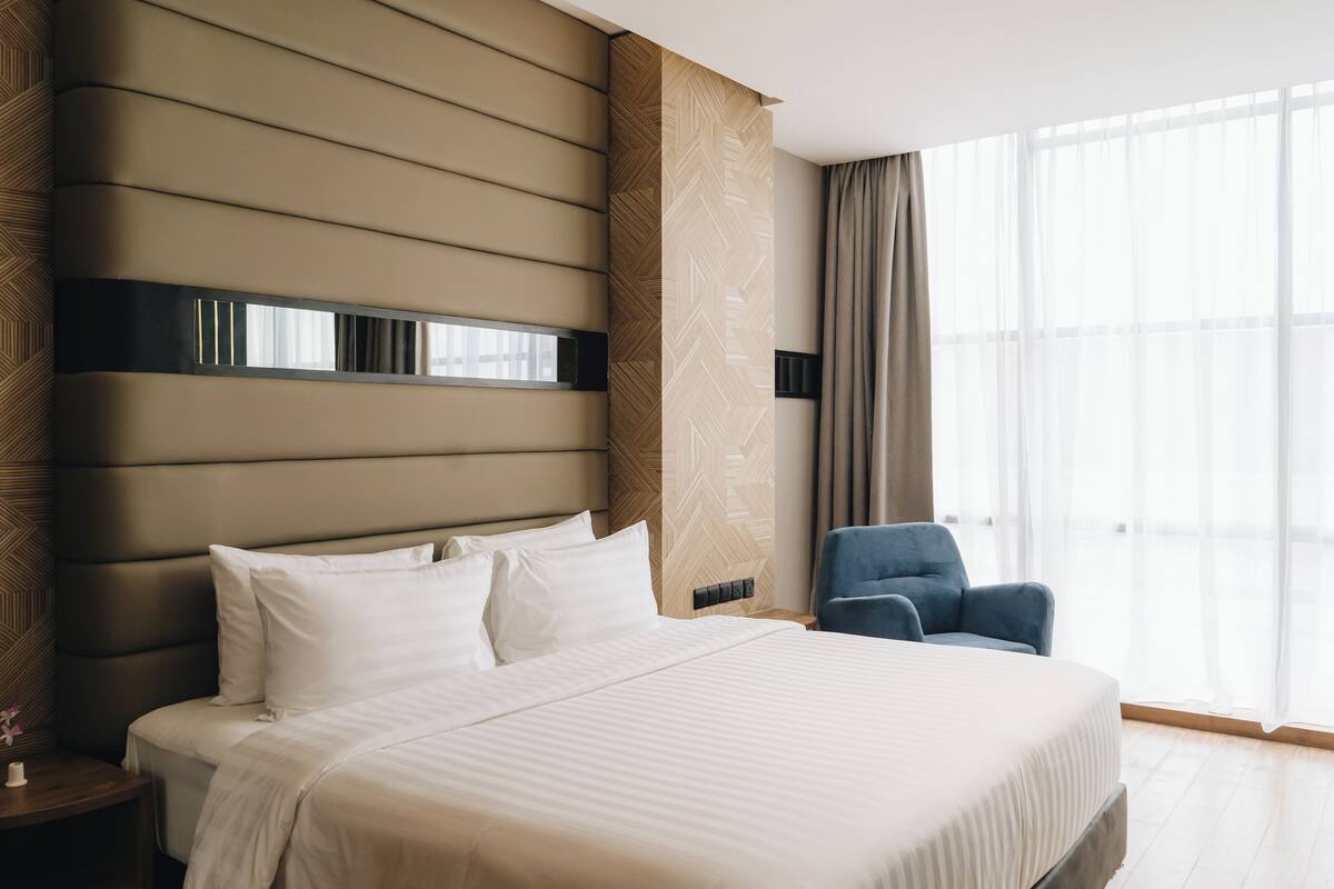 Une chambre d’hôtel moderne avec un grand lit, une table de chevet, un fauteuil bleu et une grande fenêtre avec des rideaux.