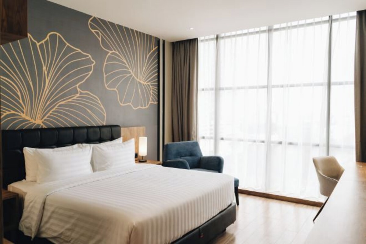 Una habitación de hotel moderna con una cama grande, un escritorio con lámpara, una silla y un amplio ventanal con cortinas.