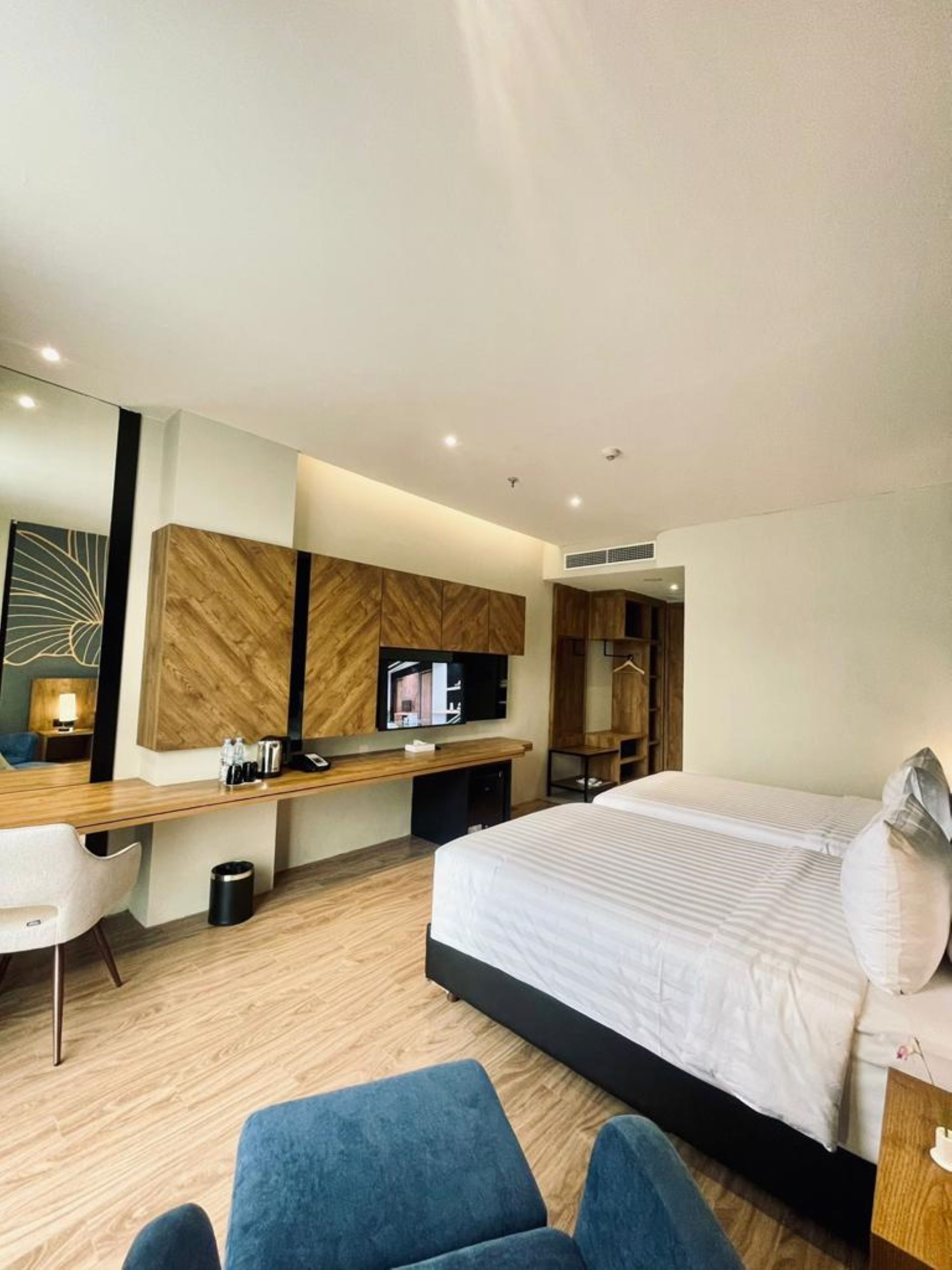 Una habitación de hotel moderna con una cama grande, un escritorio con televisor de pantalla plana, un espejo y una zona de estar con una silla azul.