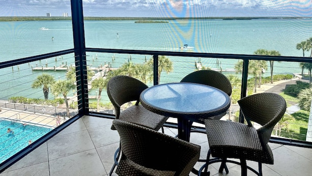 Ein Balkon mit Korbstühlen und einem Glastisch mit Blick auf einen Pool und eine Meerlandschaft, einschließlich Booten.