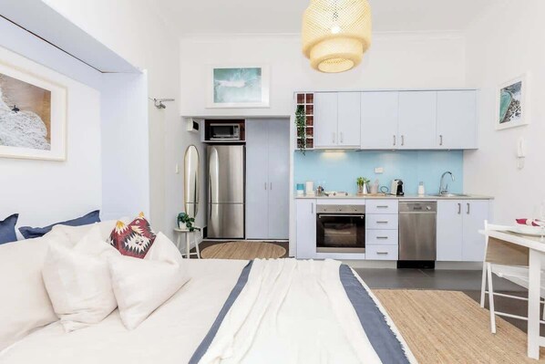 Een moderne slaapkamer met een bed, een nachtkastje en een nachttafeltje. Grenzend aan de slaapkamer bevindt zich een kleine kitchenette met een koelkast, magnetron, oven en spoelbak.