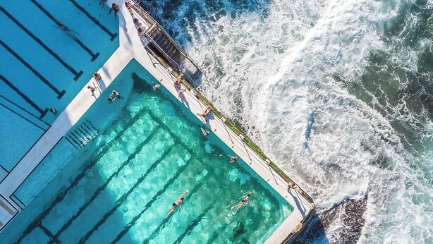 An aerial view of a swimming pool adjacent to a rocky coastline with waves crashing onto the shore. - Cozy beachfront studio with aircon (North Bondi)