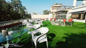 An outdoor seating area with round tables and white chairs, surrounded by green artificial grass. - Heterosis (Udaipur)