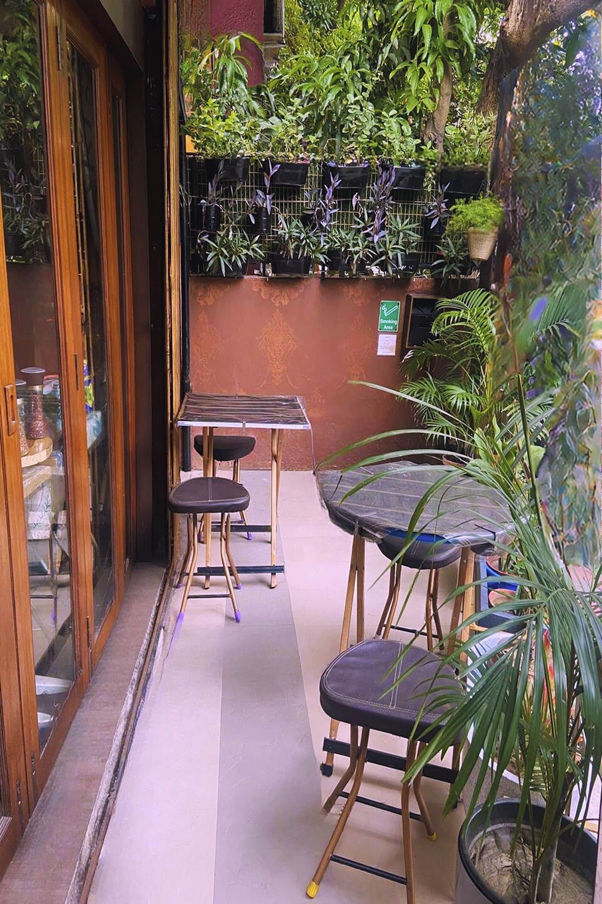 Un pequeño espacio al aire libre con una mesa y dos sillas, rodeado de plantas en macetas y una puerta de vidrio.