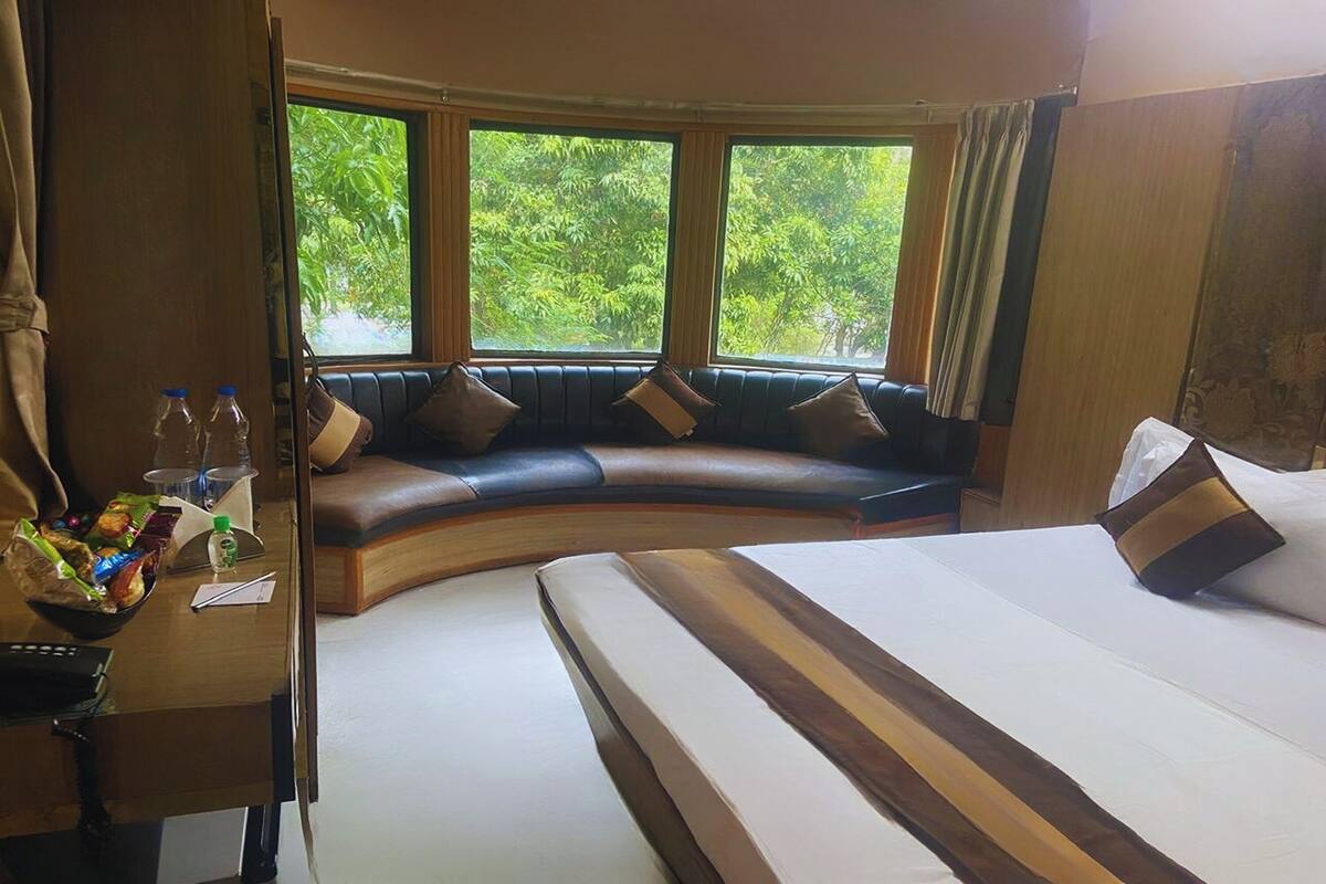 A room with a curved bench, a bed, a desk with a lamp, and large windows with a view of greenery.