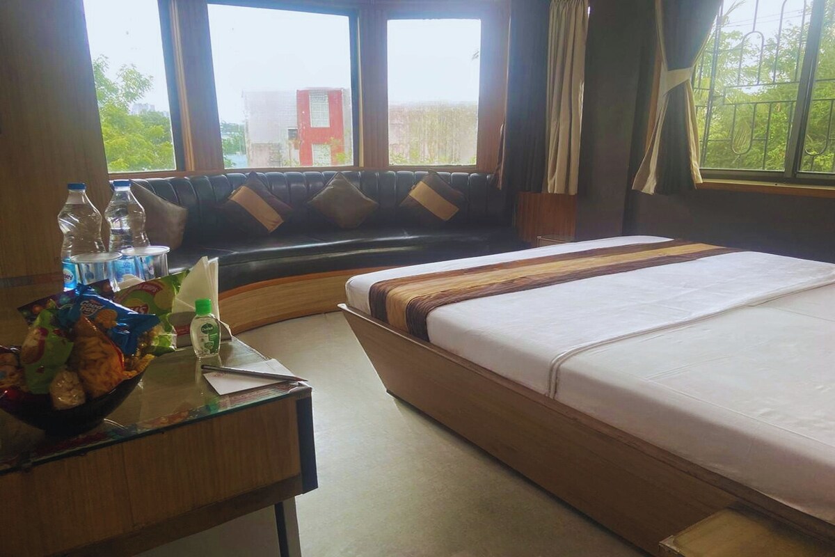 Ein Hotelzimmer mit Bett, einem Glastisch mit Snacks, einem Fenster mit Blick auf Gebäude und einer Bank mit Kissen.