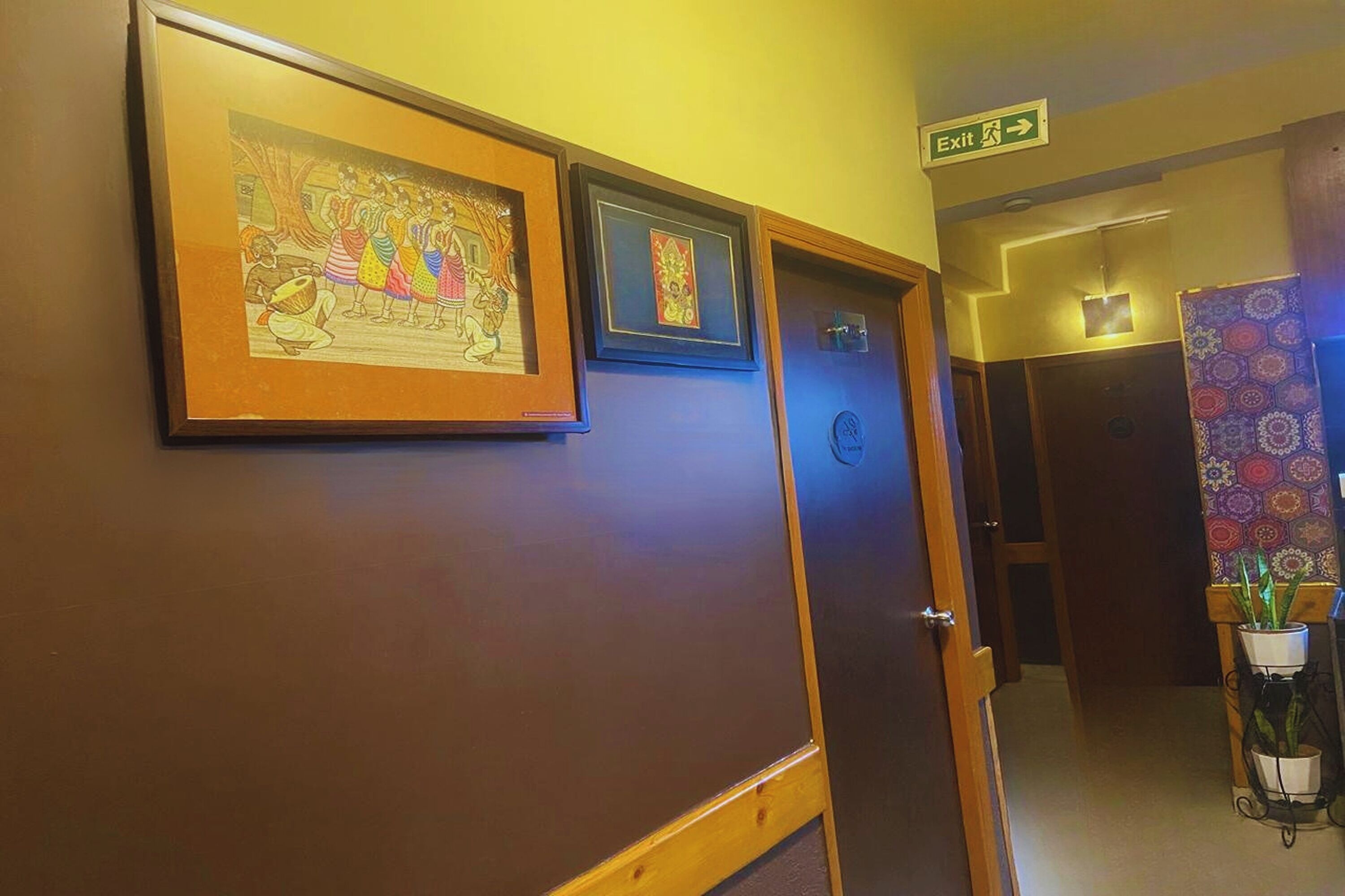 A hallway with framed artwork on the wall, an exit sign, and a door leading to another room.