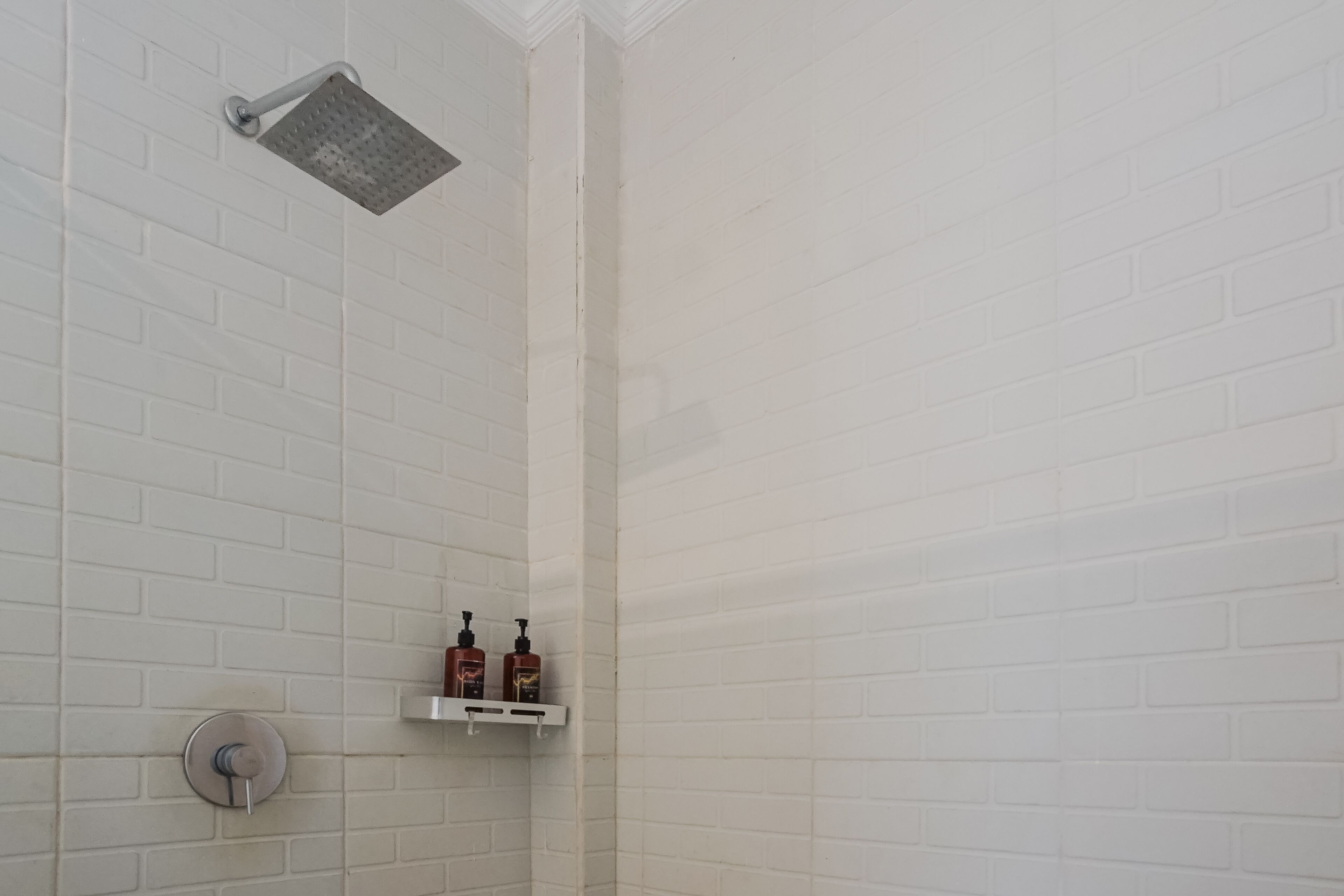 Zona de ducha con paredes de azulejos blancos, un cabezal de ducha y dos botellas de champú en un estante.