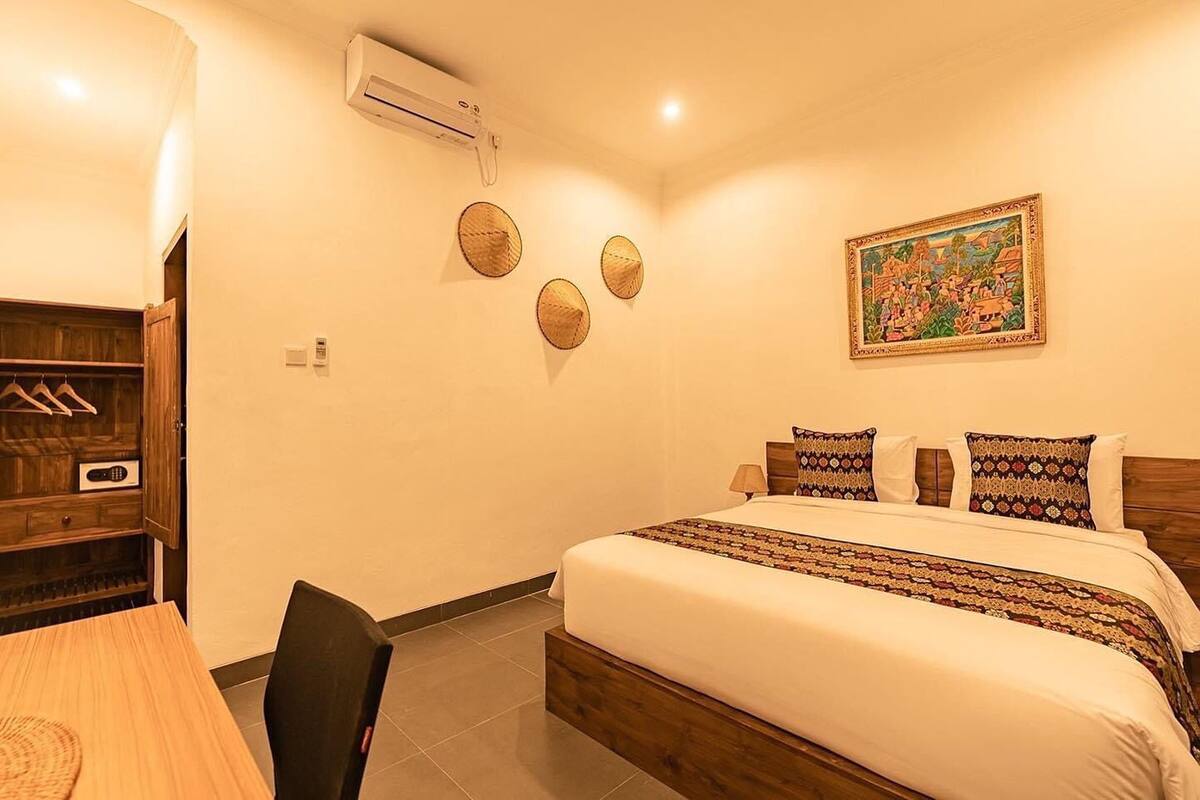 A bedroom with a bed, a wooden headboard, a wall-mounted air conditioner, a framed picture, and a wooden cabinet with hanging clothes.