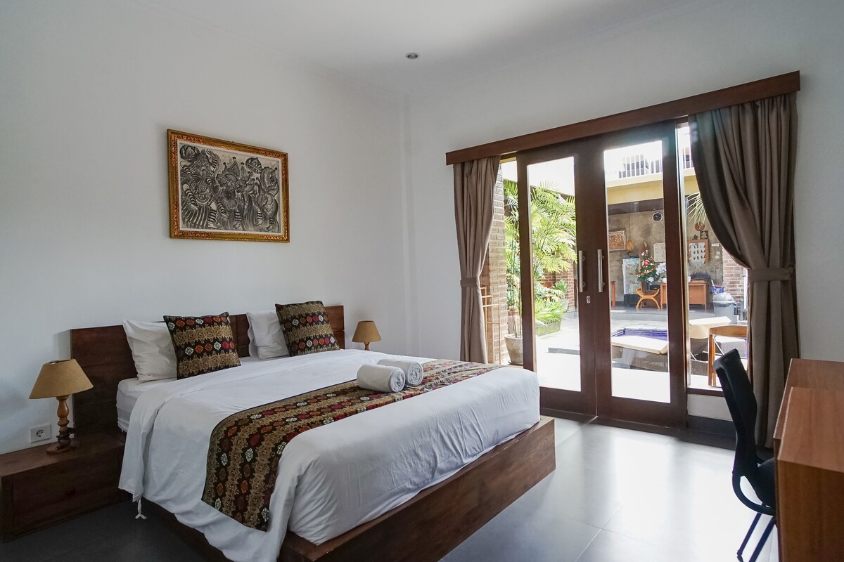 A hotel room with a bed, a desk, a chair, a lamp, a picture on the wall, and a sliding glass door leading to an outdoor area.