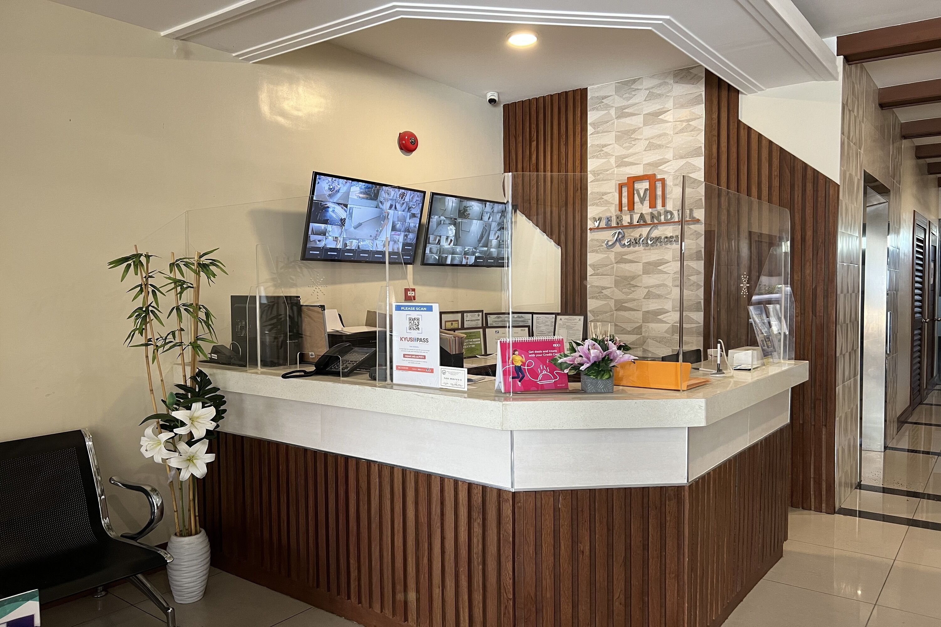 A reception desk with a modern design, featuring a wooden panel and a glass partition. There are two monitors above the desk displaying security footage. A sign indicates a QR code for KyushuPass.