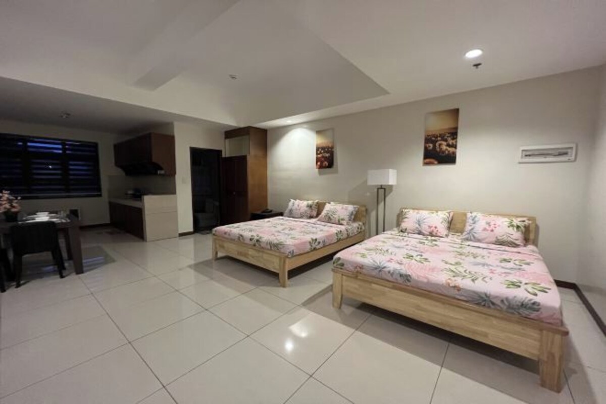 A hotel room with two wooden beds, a dining table, a kitchen area, and a window with blinds.