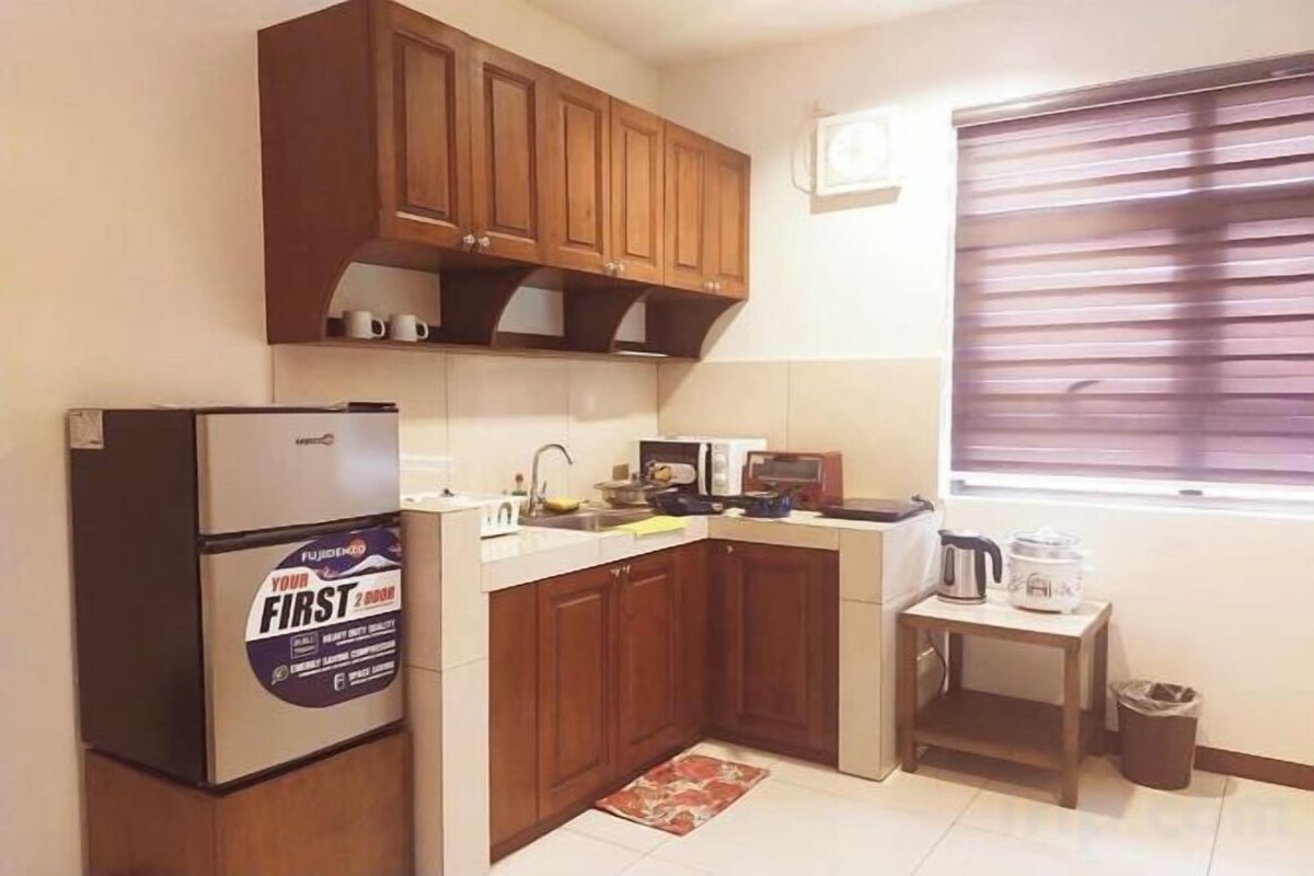 A compact kitchen with wooden cabinets, a refrigerator, a microwave, and a small table with a kettle.