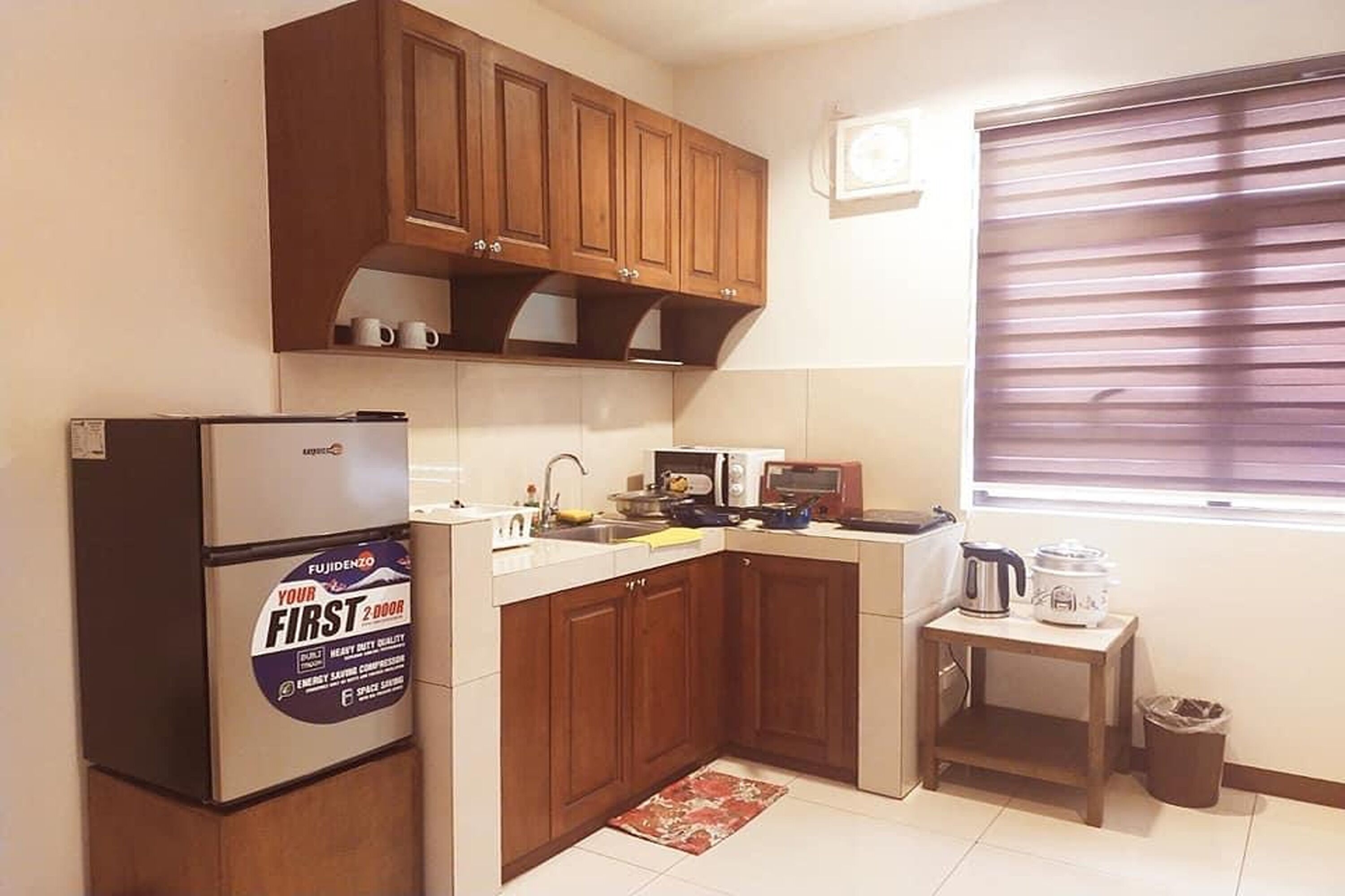 A compact kitchen with wooden cabinets, a refrigerator, a microwave, a sink, and a small table with a kettle.