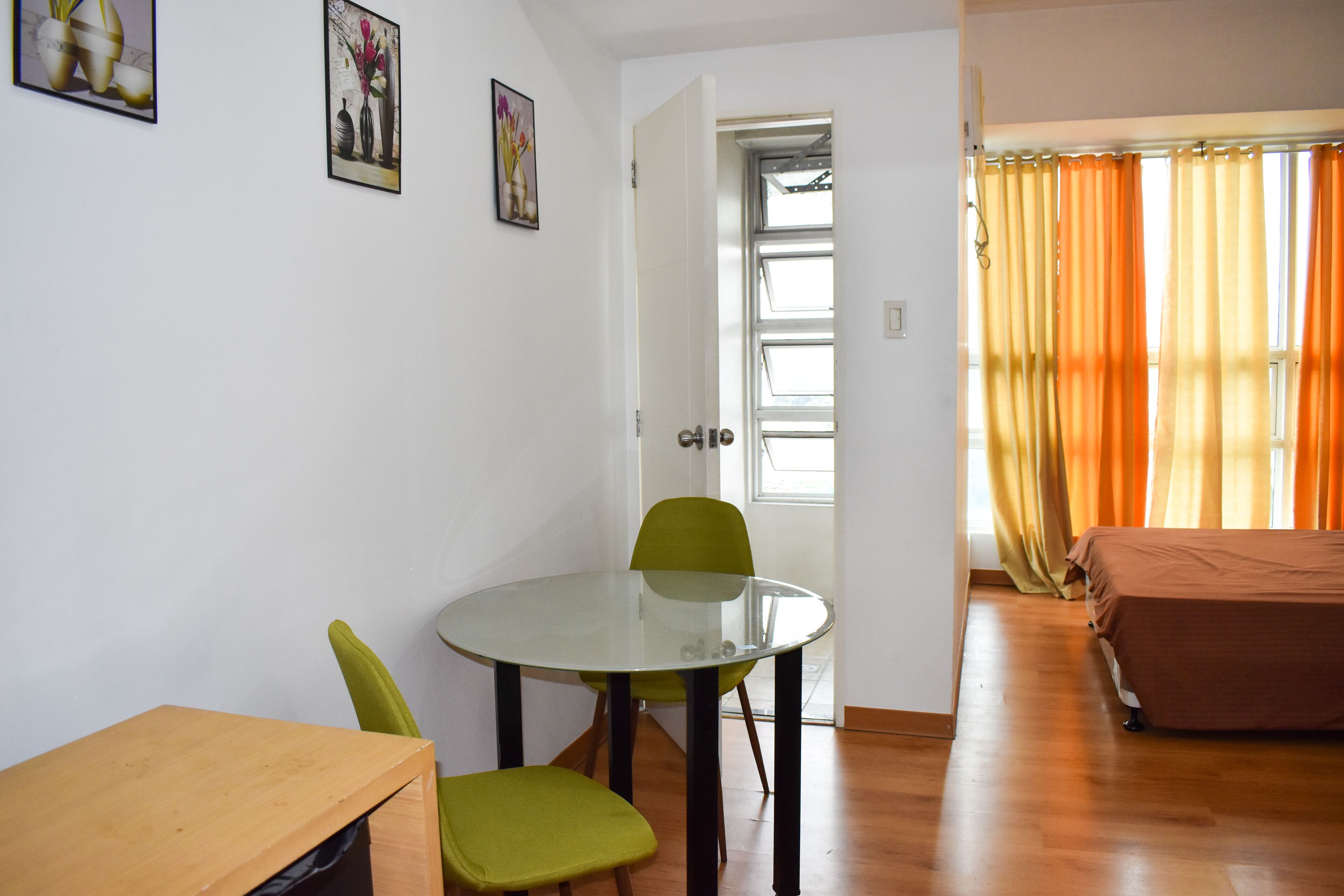 Une pièce comprenant une table avec un plateau en verre, deux chaises, un lit et une porte donnant sur une autre pièce.