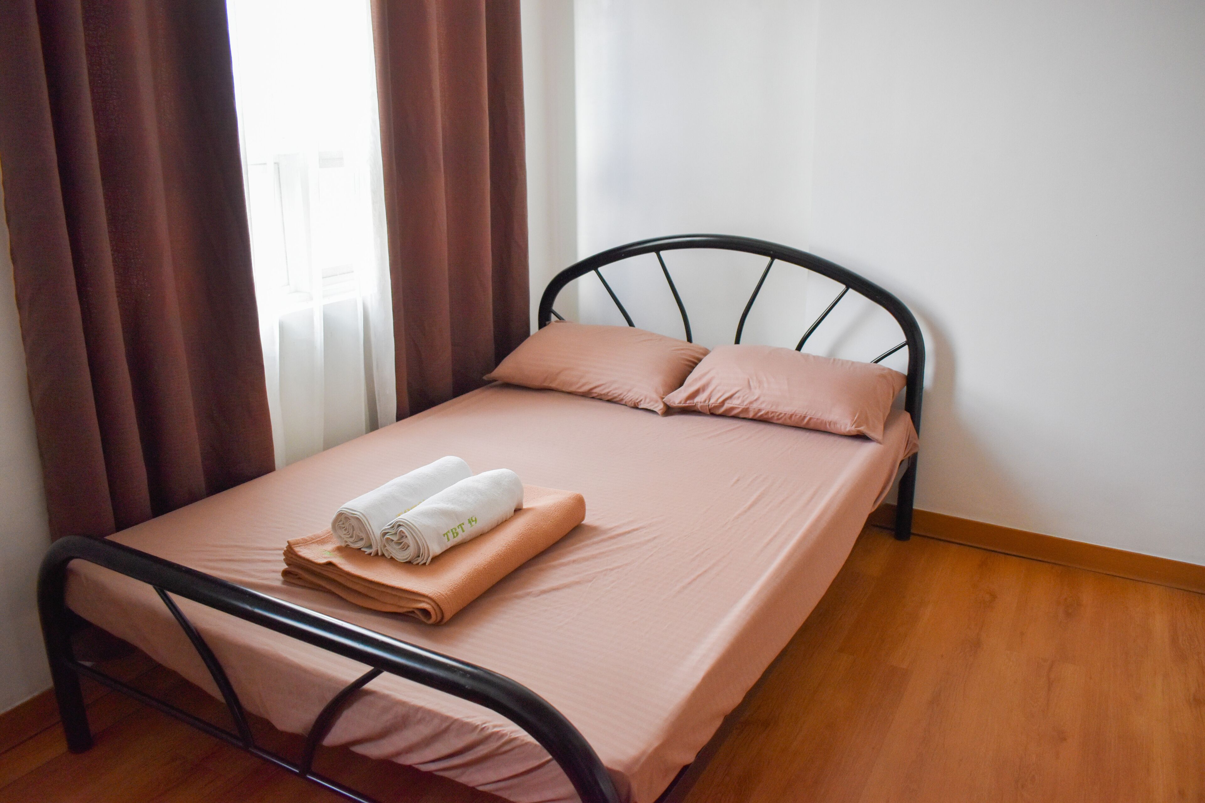 A single bed with a metal frame, a pink mattress, and two pillows. There are brown curtains partially drawn, and a window is visible in the background.