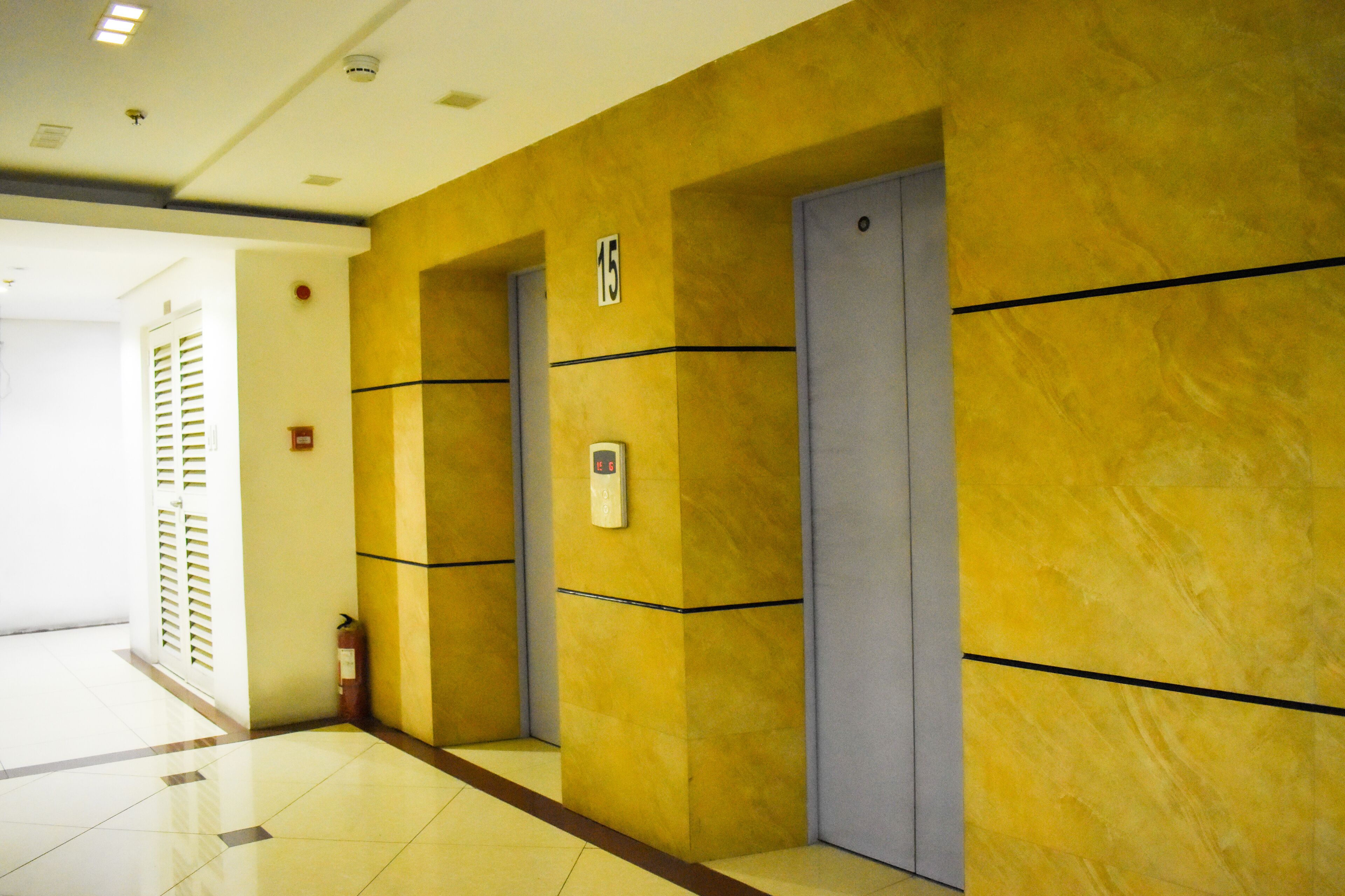 A hotel corridor with yellow walls, a fire extinguisher, and a digital elevator control panel.