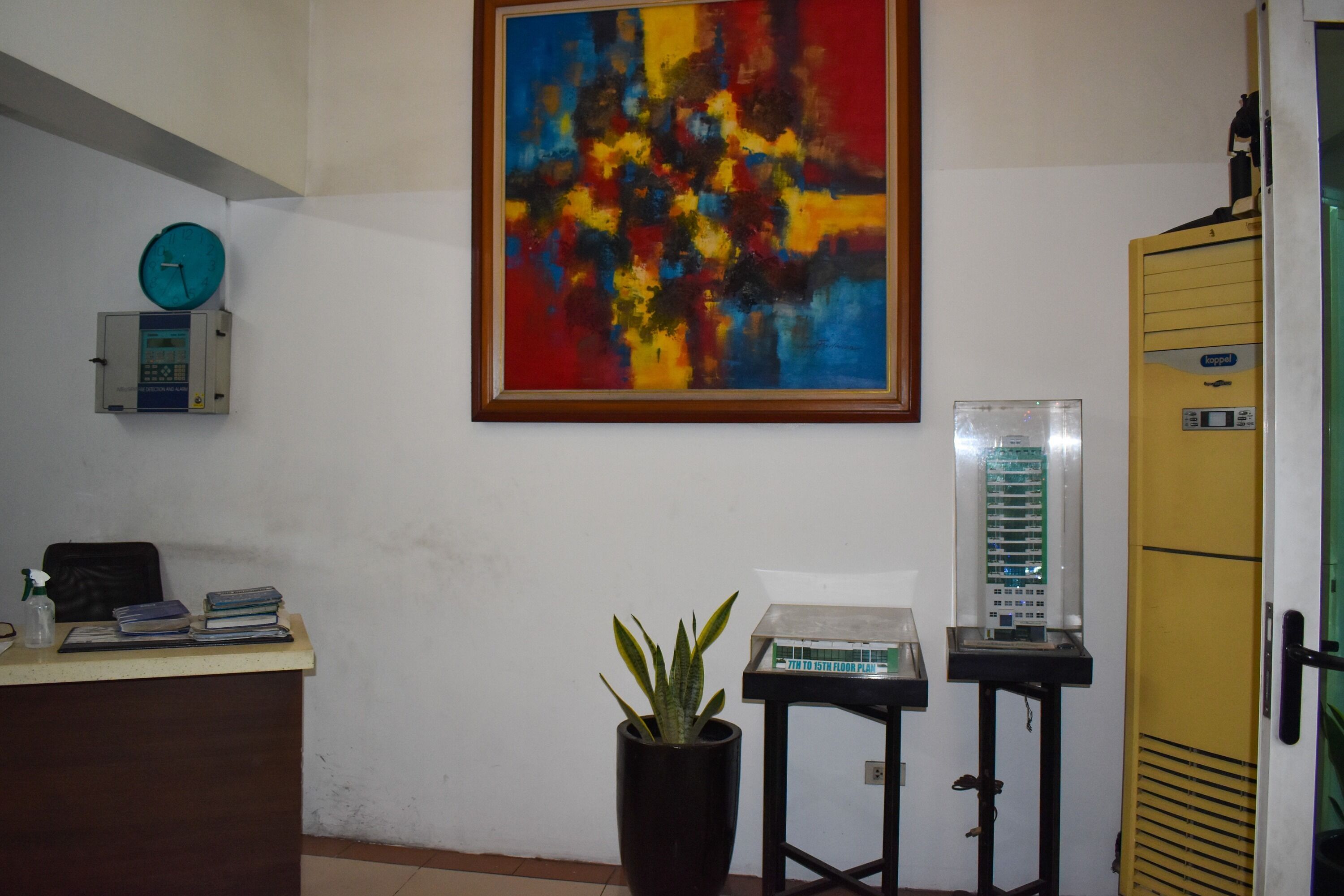 A reception area with a desk, a clock, a colorful abstract painting, a potted plant, and a filing cabinet.