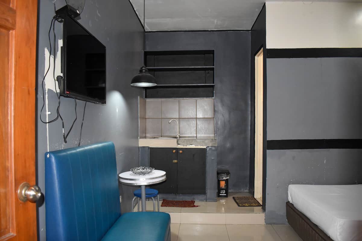 A compact room with a kitchenette, a single bed, a blue cushioned bench, and a small round table with a chair.