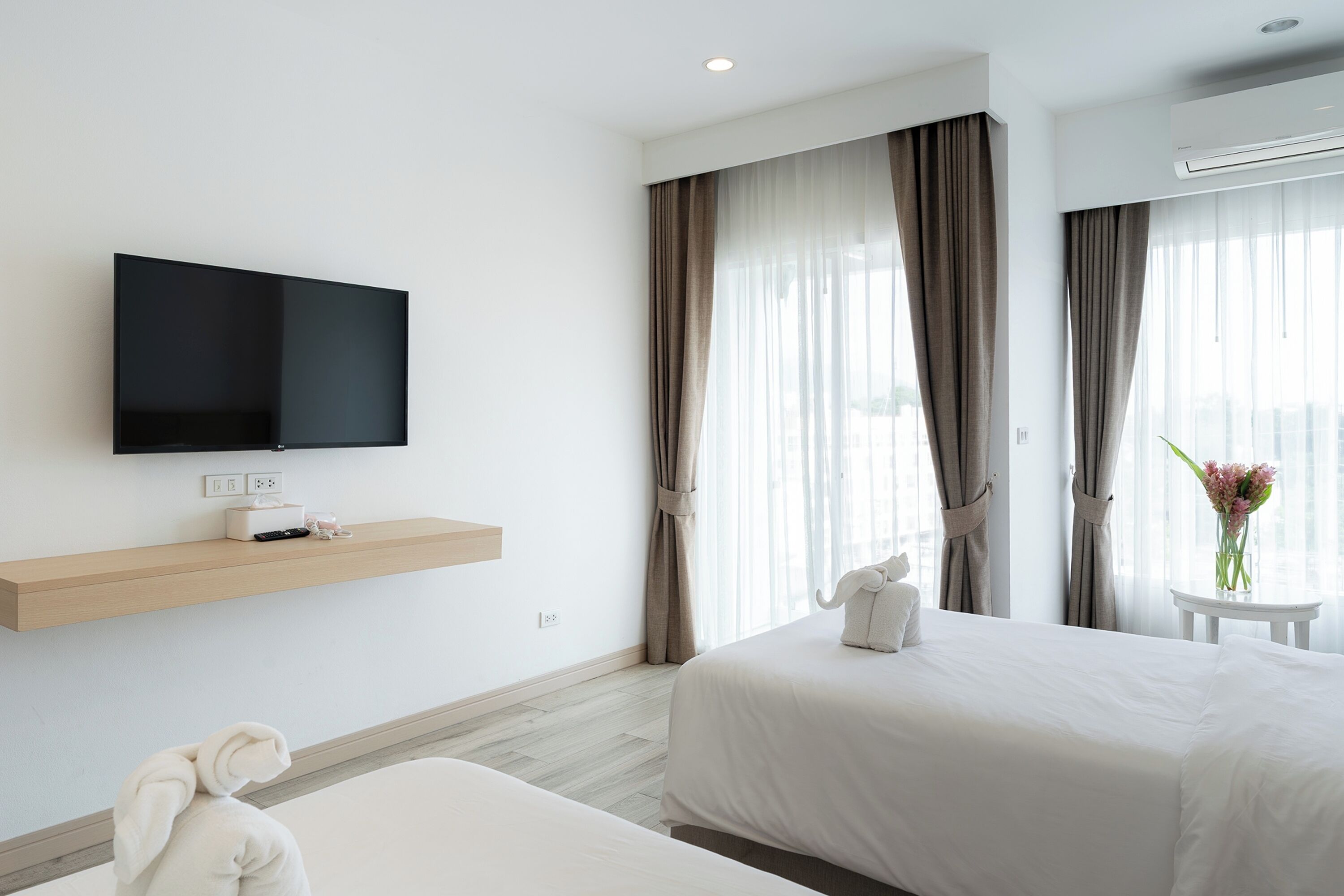 Habitación de hotel con televisión de pantalla plana montada en la pared, una mesita de noche con un florero y una cama con manta blanca.