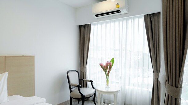 Une chambre d’hôtel avec un lit, une chaise, une petite table ornée d’un vase de fleurs et une fenêtre avec des rideaux.