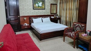 Hotelkamer met een bed, een rode bank, een houten stoel, een nachtkastje, een bijzettafeltje, een spiegel en een raam met gordijnen.