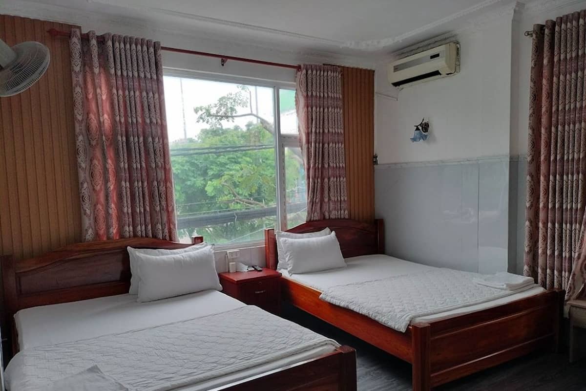 Une chambre d’hôtel avec deux lits en bois, une grande fenêtre avec des rideaux, un appareil de climatisation et une petite table avec une chaise.