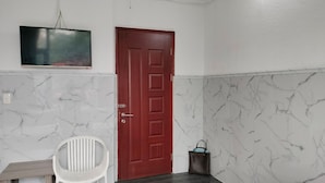 Een kamer met een wand met een marmerpatroon, een houten deur, een witte stoel, een klein tafeltje, een aan de muur bevestigde televisie, en een donkere houten vloer.