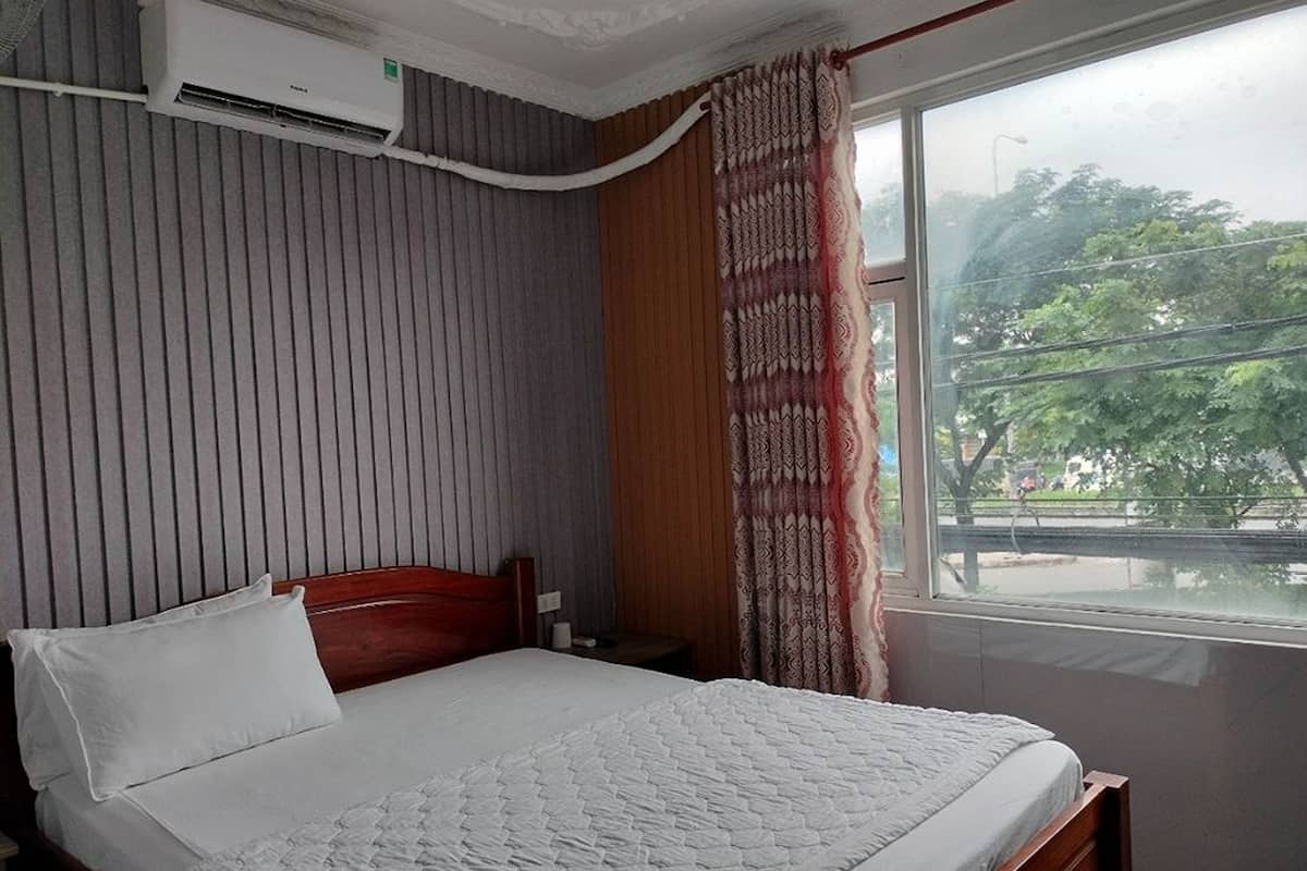 Une chambre d’hôtel avec un lit, une fenêtre avec des rideaux, un climatiseur fixé au mur, et une vue sur des arbres et une rue.