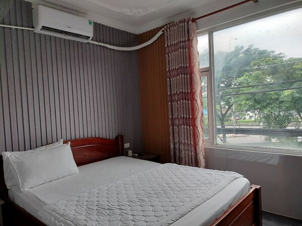 Une chambre d’hôtel avec un lit, une fenêtre avec des rideaux, un climatiseur fixé au mur, et une vue sur des arbres et une rue.