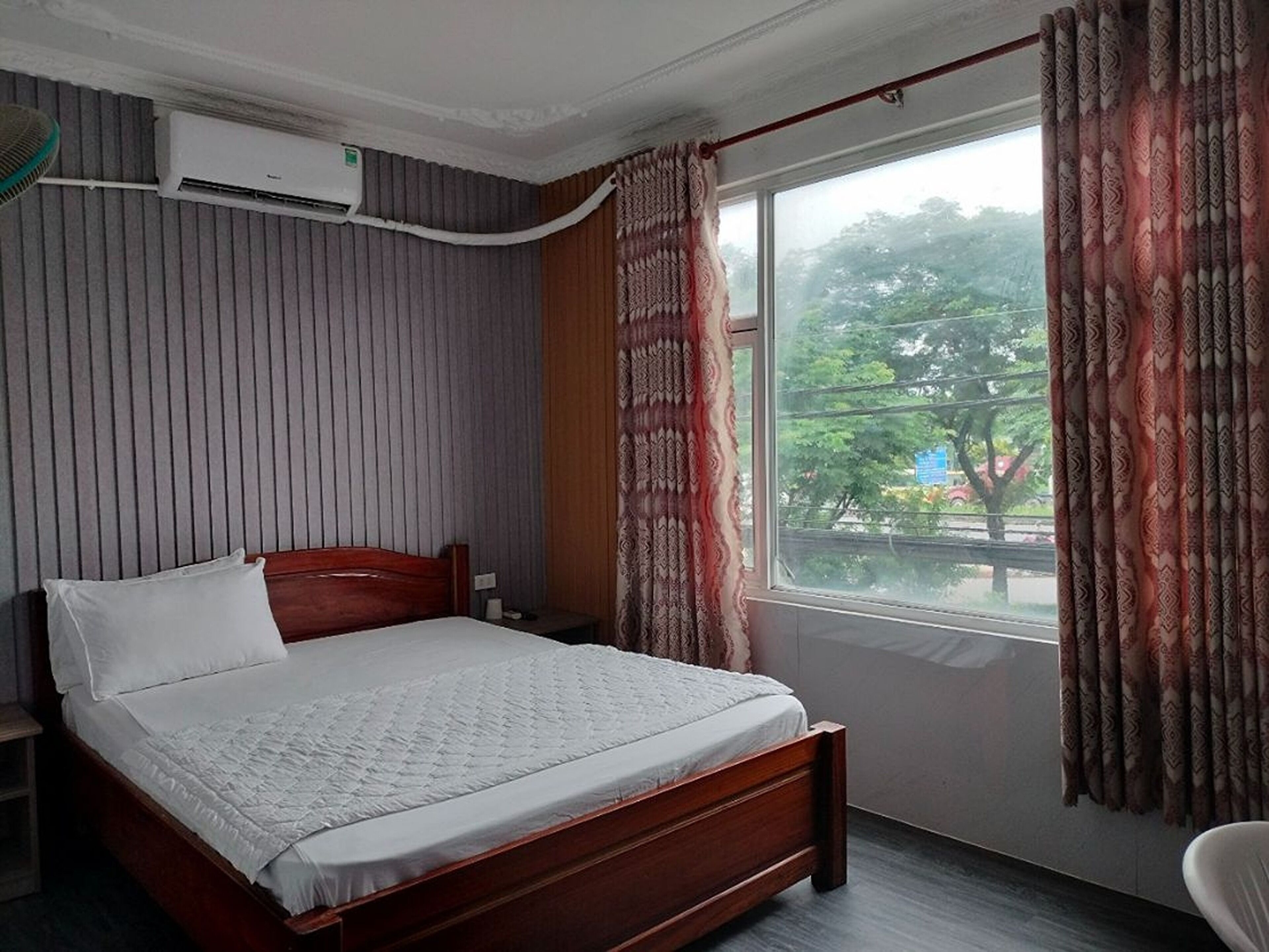 Habitación de hotel con una cama de madera, una ventana con cortinas estampadas y un equipo de aire acondicionado en la pared.