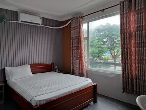 Une chambre d’hôtel avec un lit en bois, une fenêtre ornée de rideaux à motifs et un climatiseur fixé au mur.