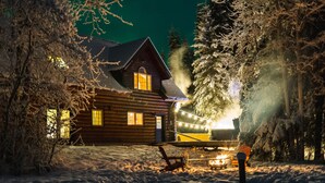 Uma cabana de tronco com um pátio coberto de neve, um alpendre com uma cadeira e uma guirlanda de luzes.