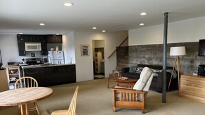 A modern living room with a wooden dining table, two chairs, a sofa, and a coffee table. A kitchen with a microwave and stove is visible in the background. - Garden apt. above the beach close to hiking trails and beach. (Stinson Beach)