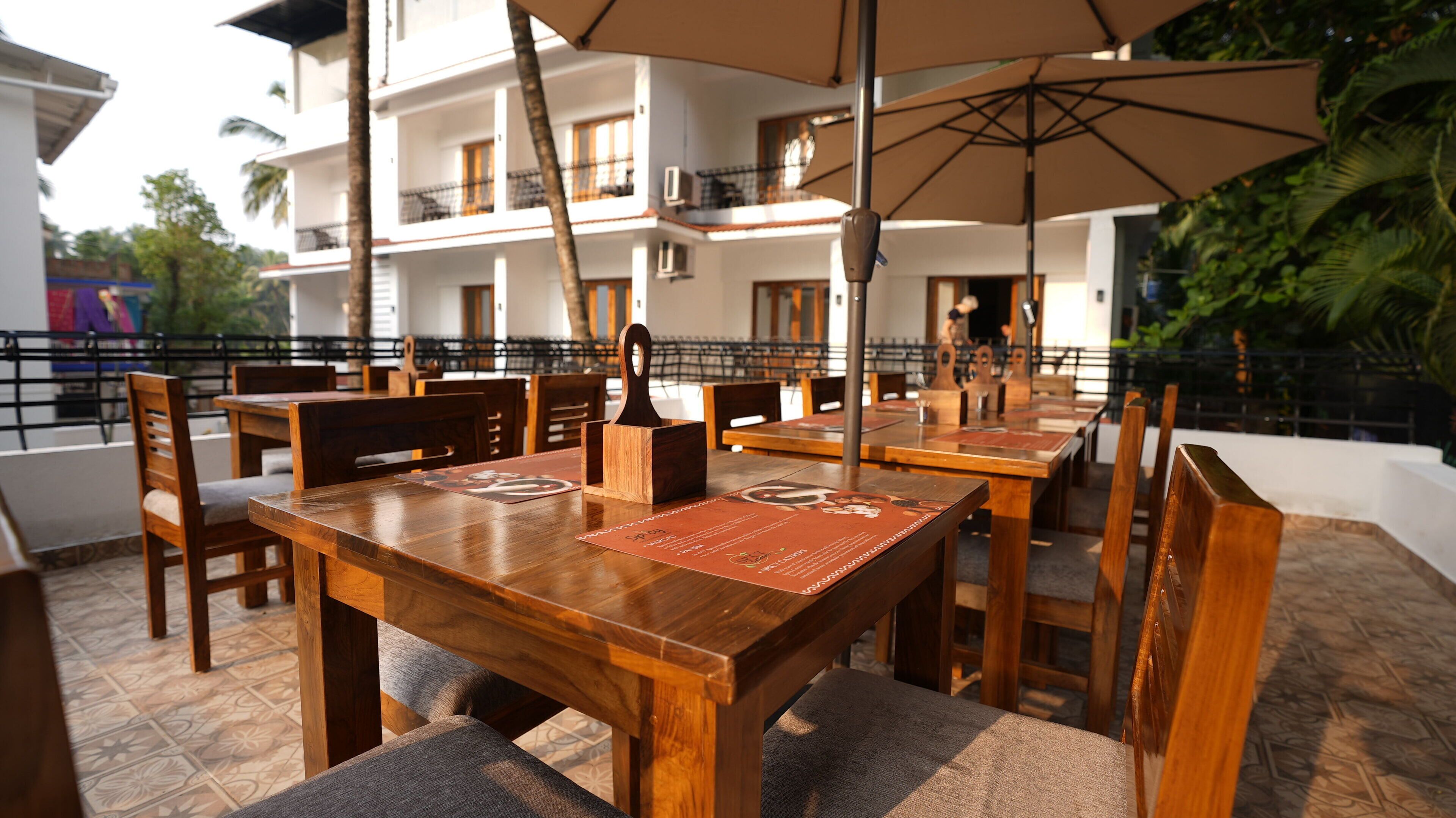 A restaurant with wooden tables and chairs, outdoor umbrellas, and a view of a white building with balconies.