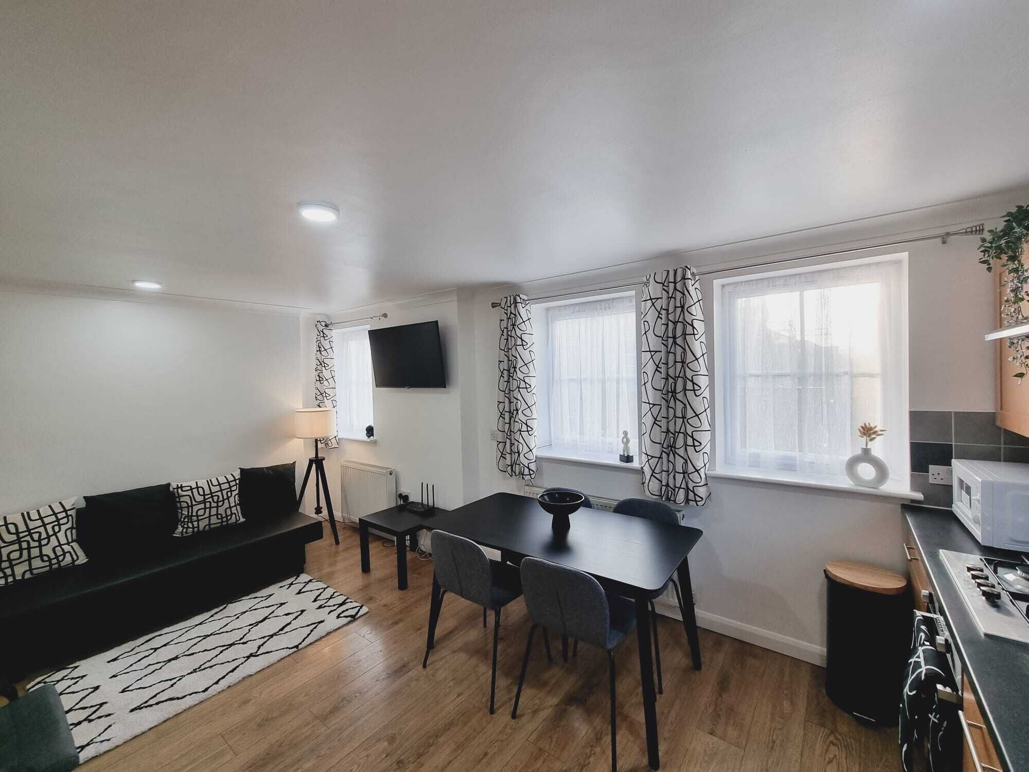 A modern living room with a black sofa, dining table, and chairs. There is a TV mounted on the wall, a lamp, and a window with patterned curtains.