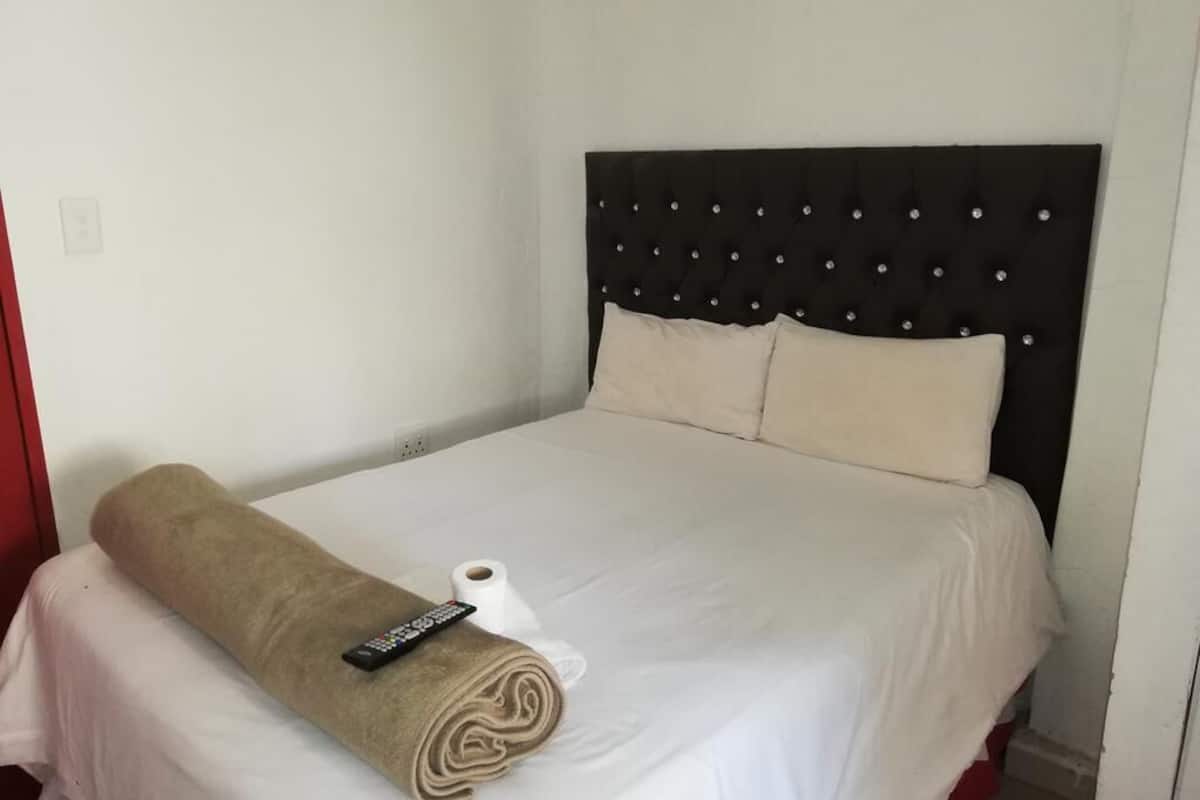 A neatly made bed with a rolled towel and remote control, a plain white wall, and a simple headboard with buttons.