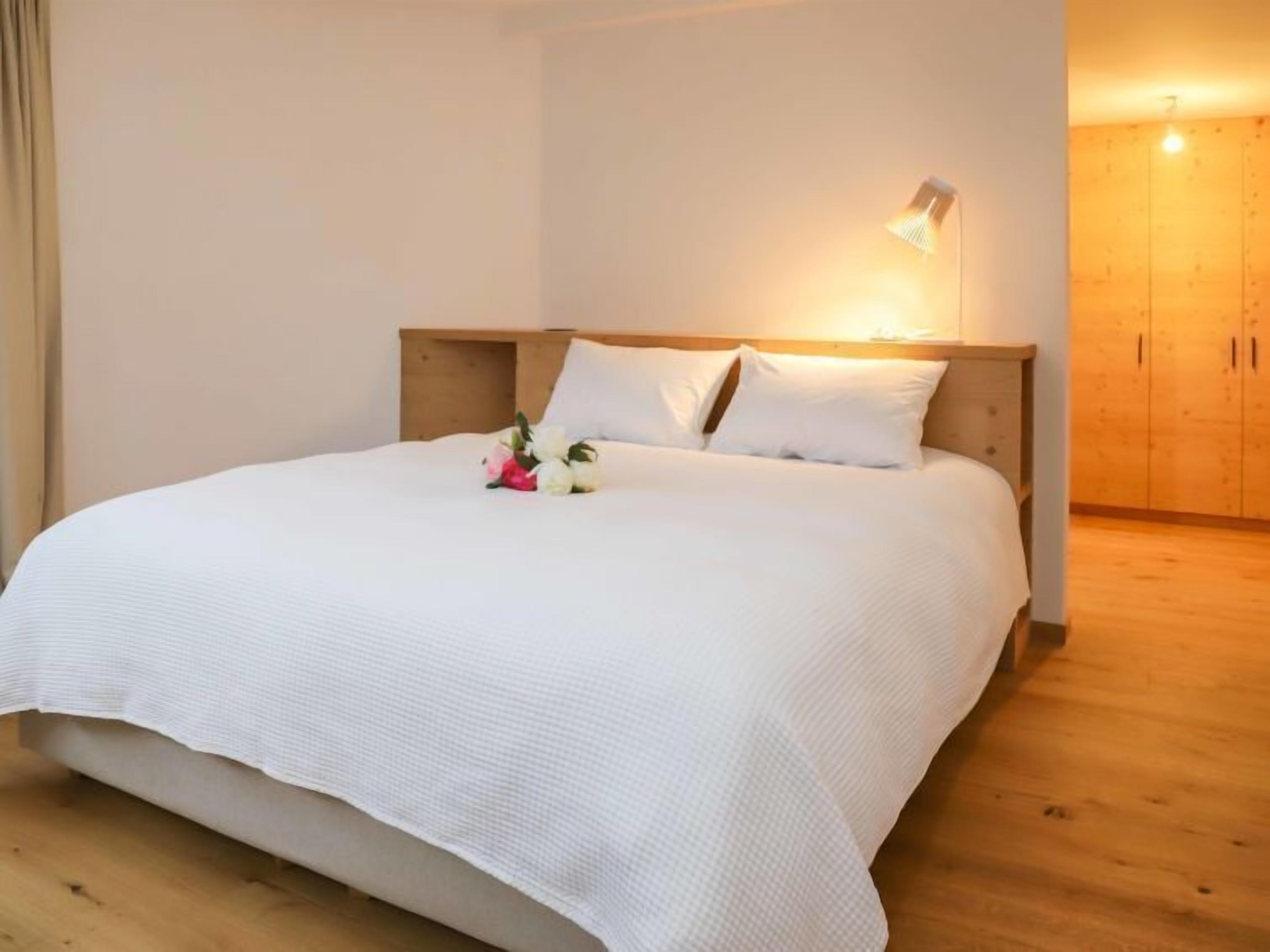 A neatly made bed with white linens and a small bouquet of flowers. There is a wooden headboard and a wall-mounted light fixture.