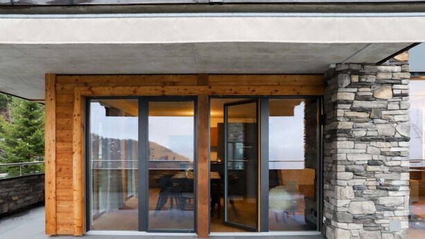 Ein moderner Gebäudeeingang mit Glastür und Holzrahmen, einer Steinsäule und Blick ins Innere.