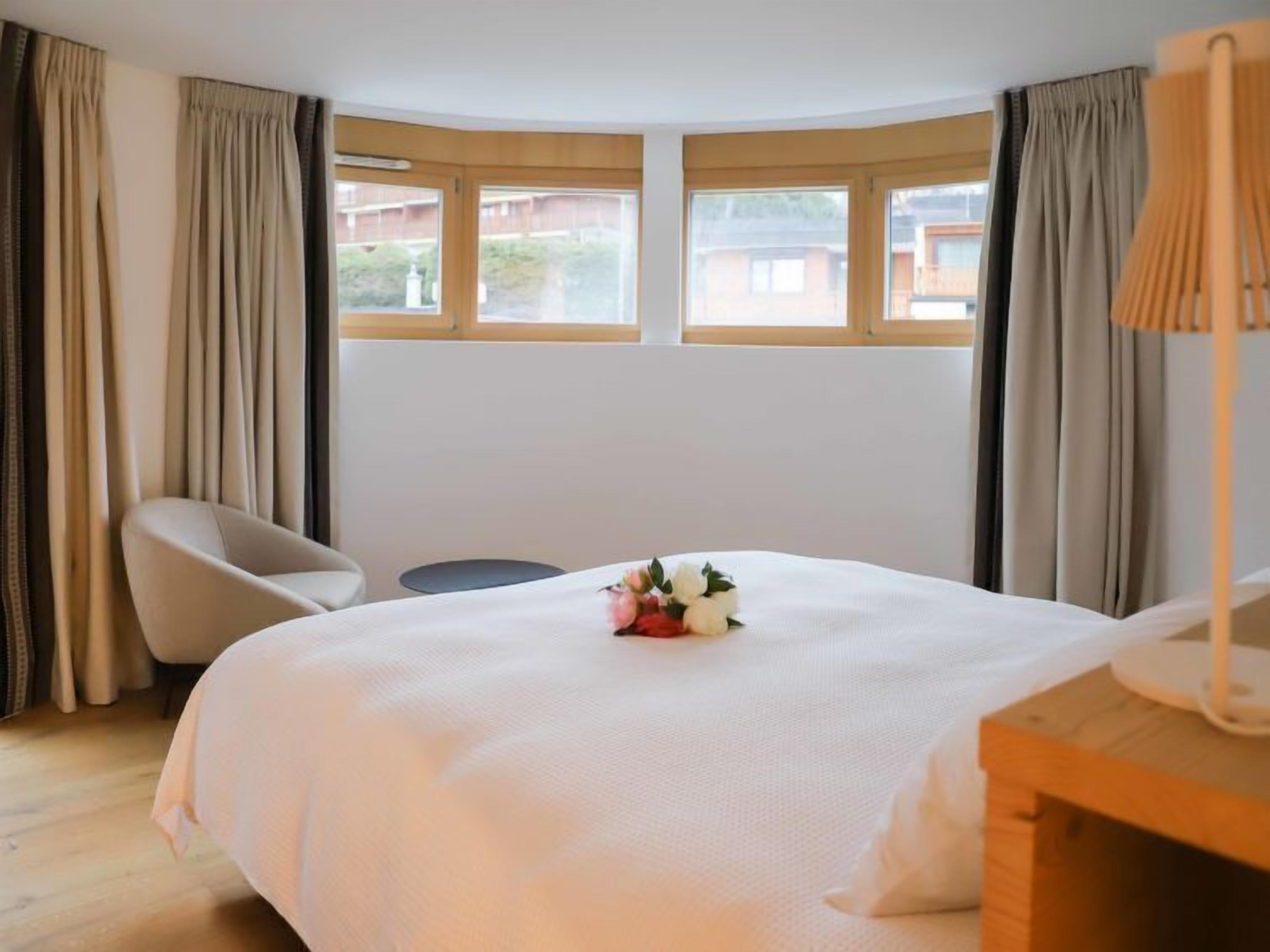 A hotel room with a large bed, a chair, a small table, and a lamp. There are two windows with curtains.