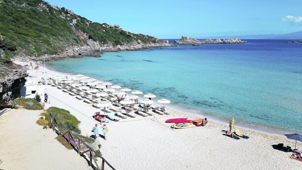 Une plage de sable blanc, avec une eau d’un bleu cristallin et des falaises rocheuses. Des parasols et des chaises longues sont disposés le long du rivage.