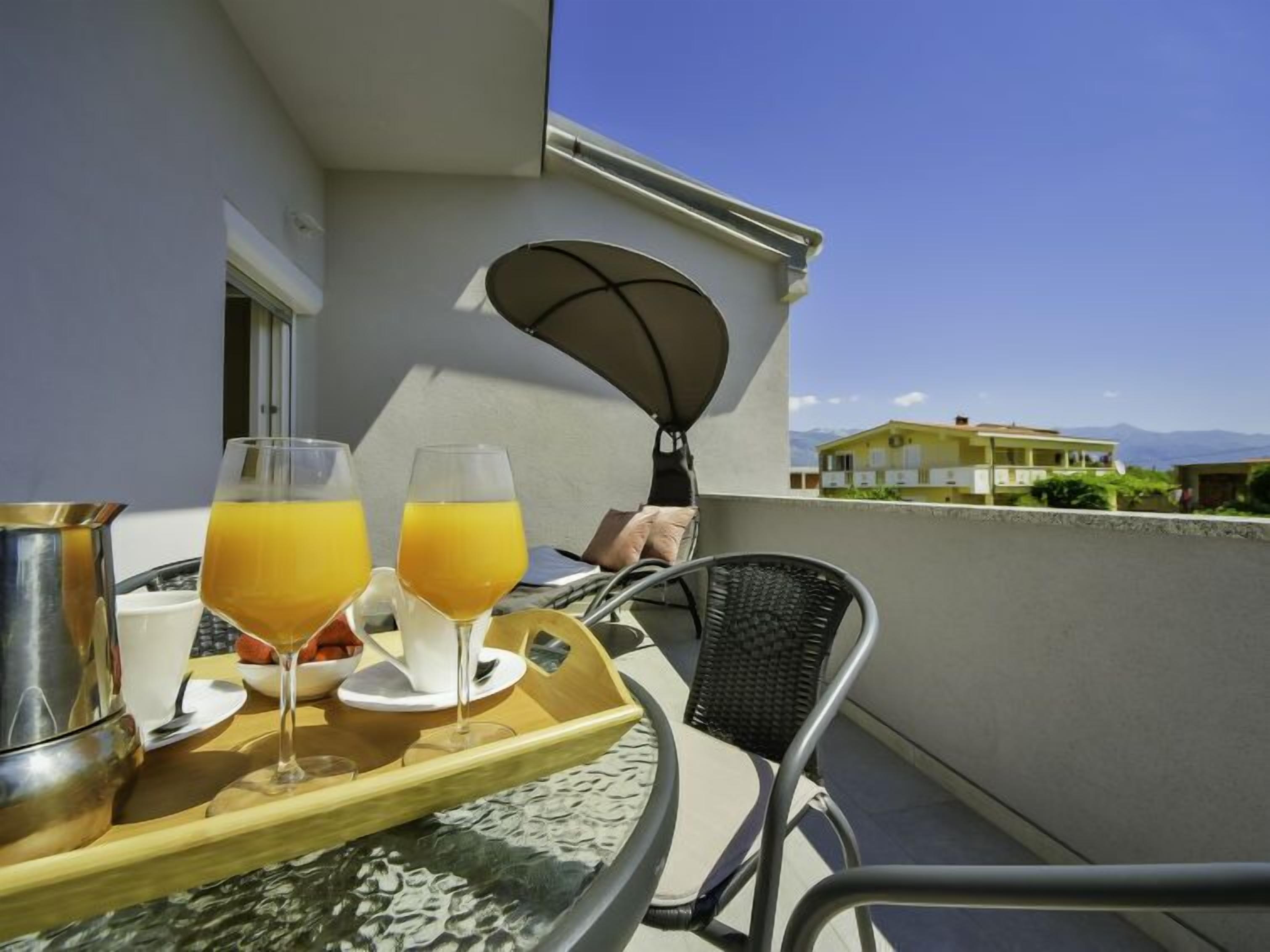 A balcony with a table set for breakfast, including two glasses of orange juice, a coffee cup, and a metal pitcher.