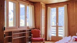 Una habitación de madera con una cama, una silla roja y un ventanal con cortinas.