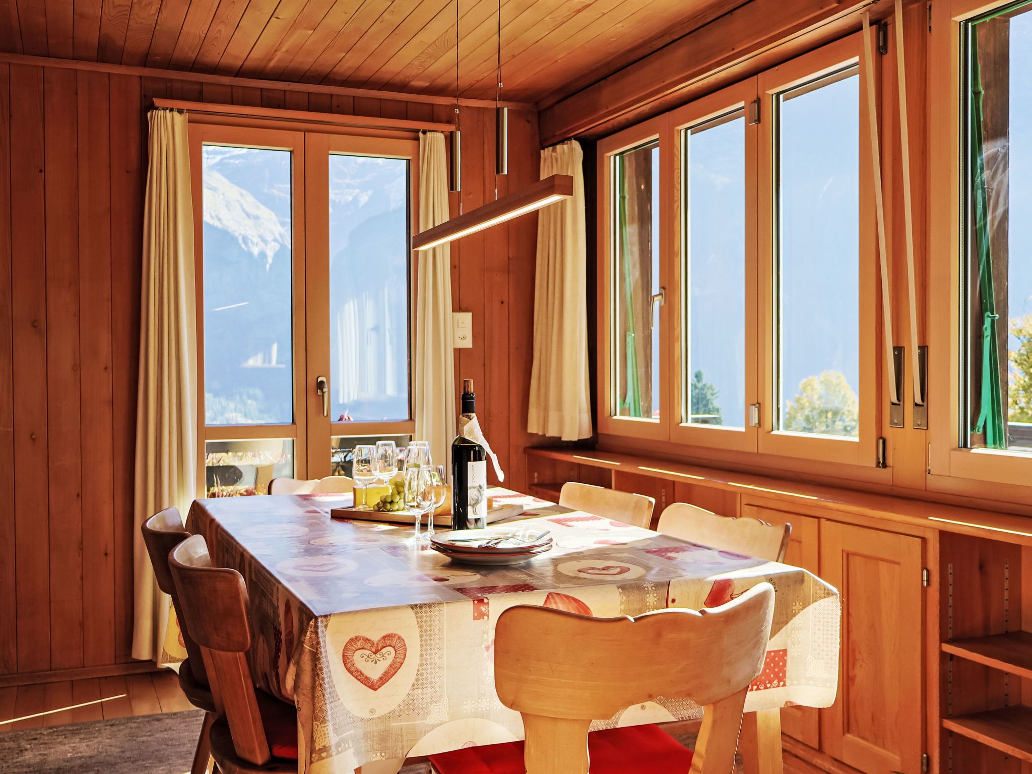Une salle à manger en bois avec une table dressée pour quatre personnes, de grandes fenêtres et une bouteille de vin posée sur la table.
